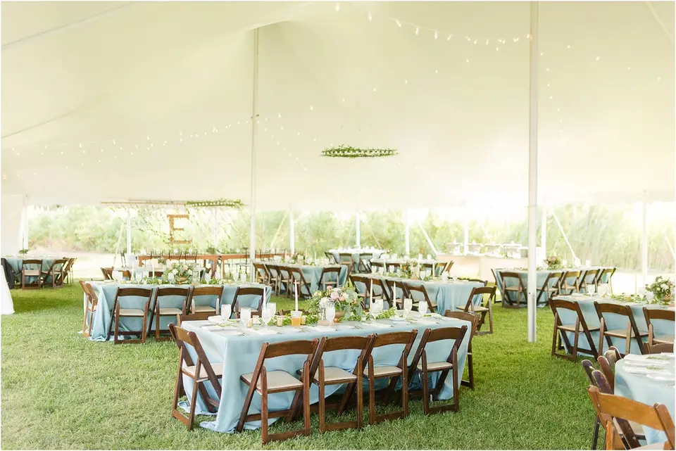 Reception tent with tables in light blue linens, wooden chairs and string lights over a grassy floor