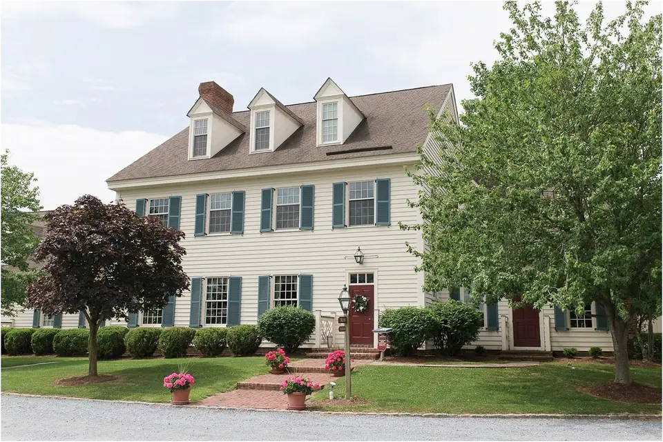 White colonial-style house with green shutters and a brick walkway, pink flower pots at the door
