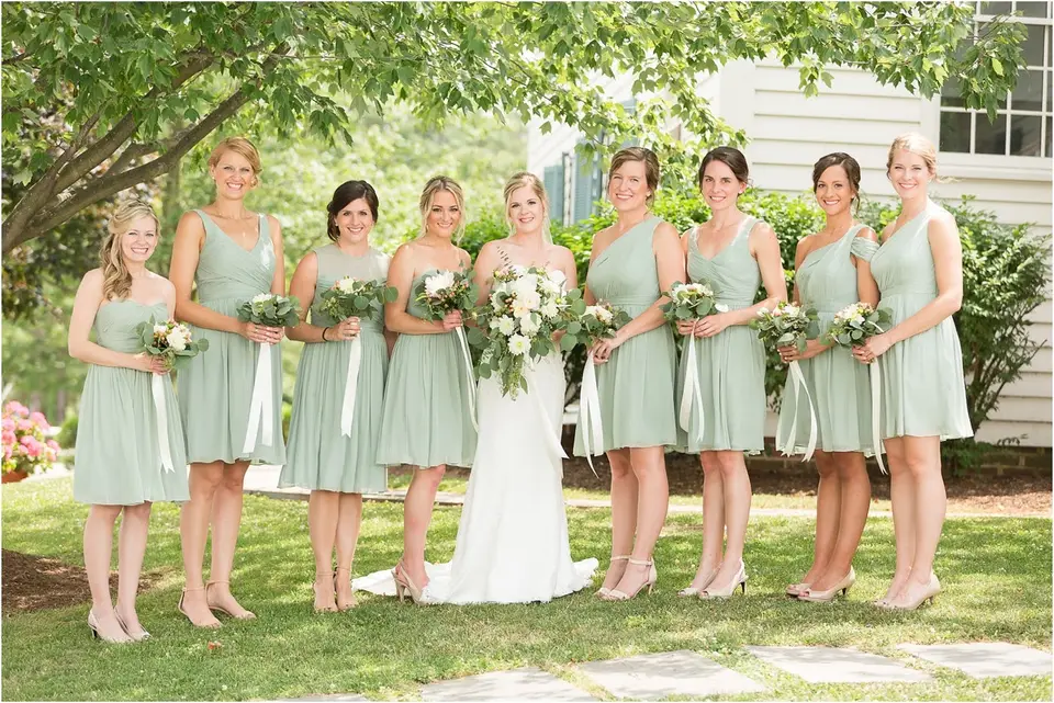 Bride standing with eight bridesmaids in sage green dresses holding bouquets outdoors