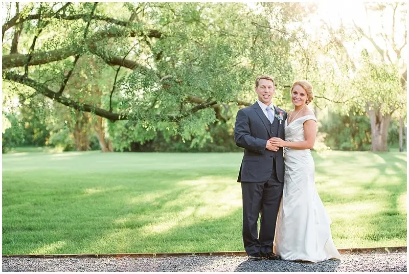 Bride and groom standing arm in arm on a lawn beneath a large tree
