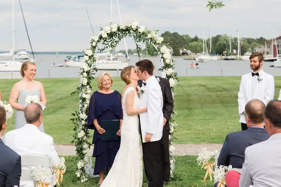 Couple kissing under a floral arch on a waterfront lawn with sailboats behind them and guests watching