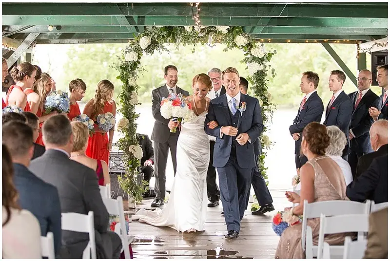 Bride and groom walking up the aisle under a floral arch on a covered dock as guests cheer