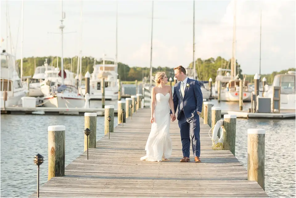 Bride and groom holding hands and walking down a wooden dock lined with posts at a marina with sailboats