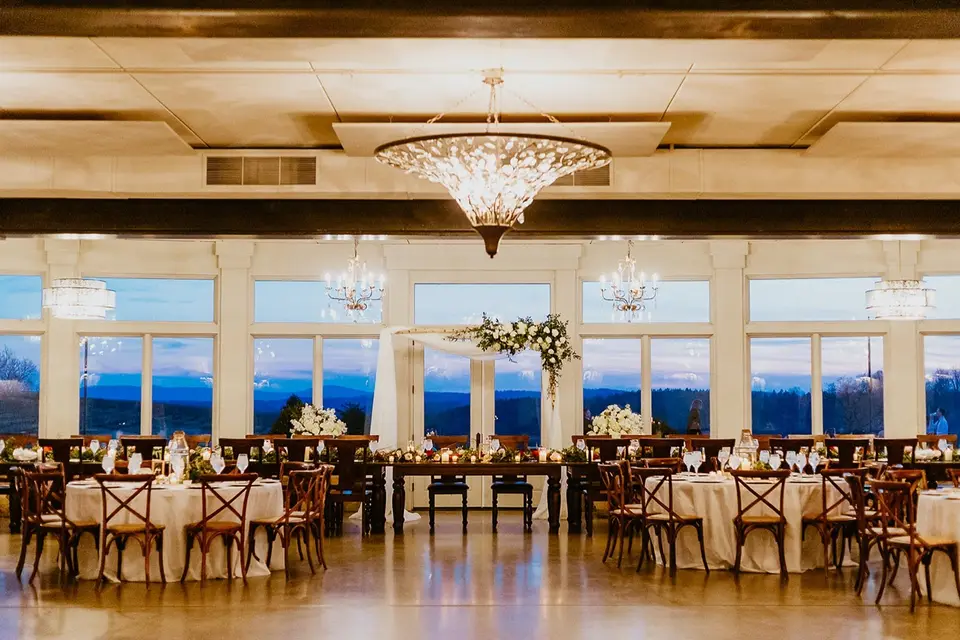 Reception room with set round tables, chandeliers and tall windows overlooking a view at dusk
