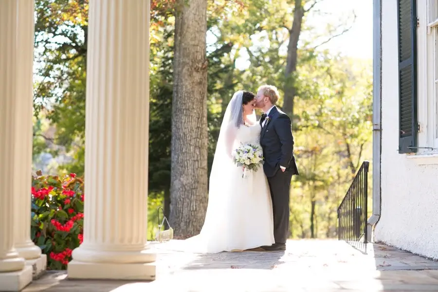 Bride and groom kissing on a stone porch beside white columns with fall foliage behind