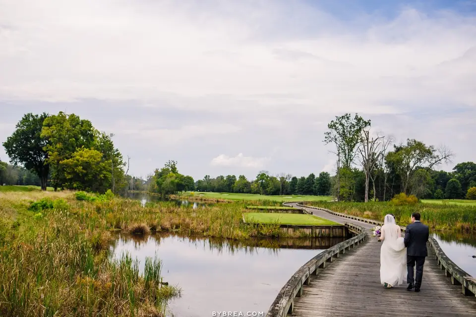 Bride and groom walking on a wooden boardwalk over a pond toward a wooded shoreline