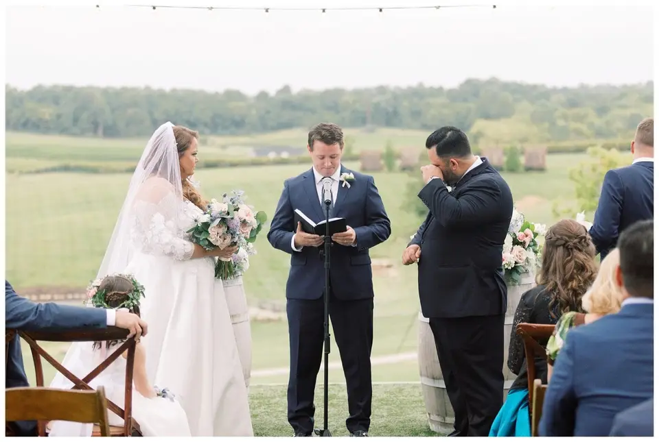 Bride and groom at an outdoor vineyard ceremony as the officiant reads and the groom wipes his face