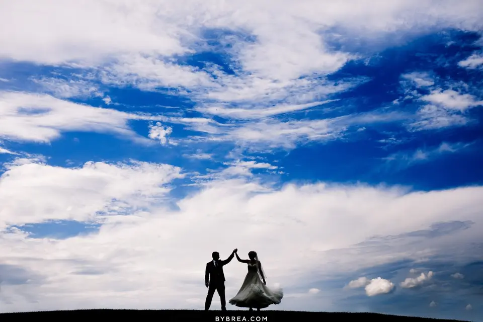 Silhouette of a bride and groom dancing on a hilltop under a large cloudy blue sky