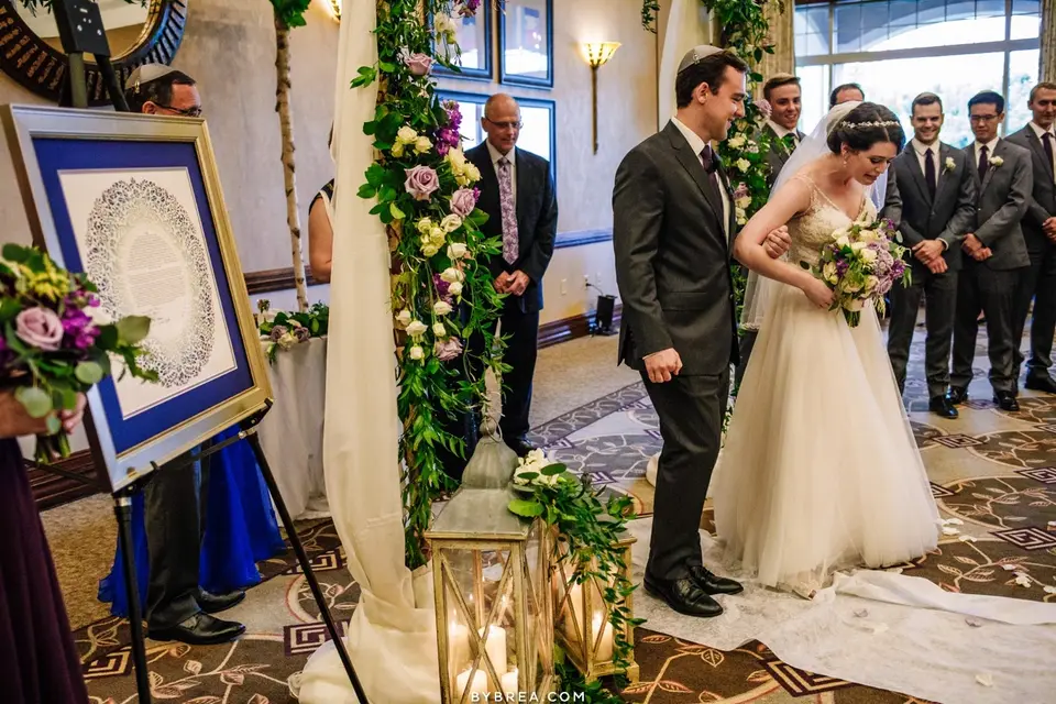 Groom in a kippah and bride standing under a floral-draped chuppah beside a framed ketubah