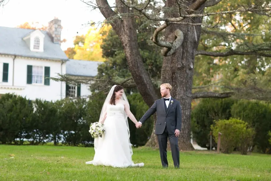 Bride and groom holding hands and walking on a lawn near a large tree and white house