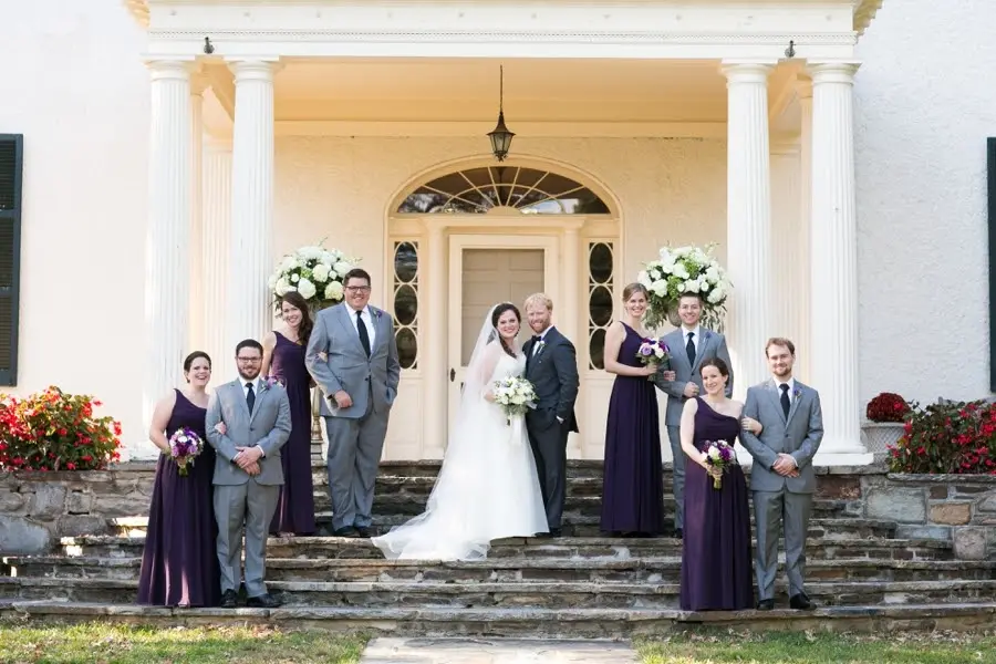 Bride, groom and bridal party in plum dresses and gray suits on porch steps of a columned house
