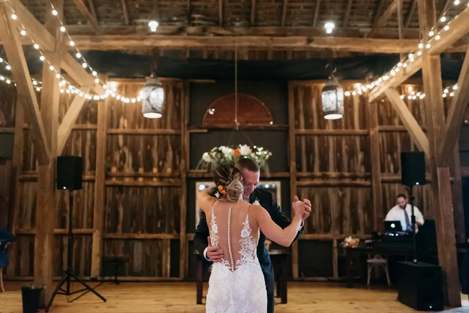 Bride and groom dancing their first dance in a rustic barn with string lights, DJ setup behind