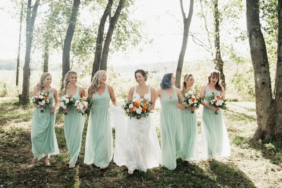 Bride walking with five bridesmaids in sage green dresses holding bouquets through a wooded area