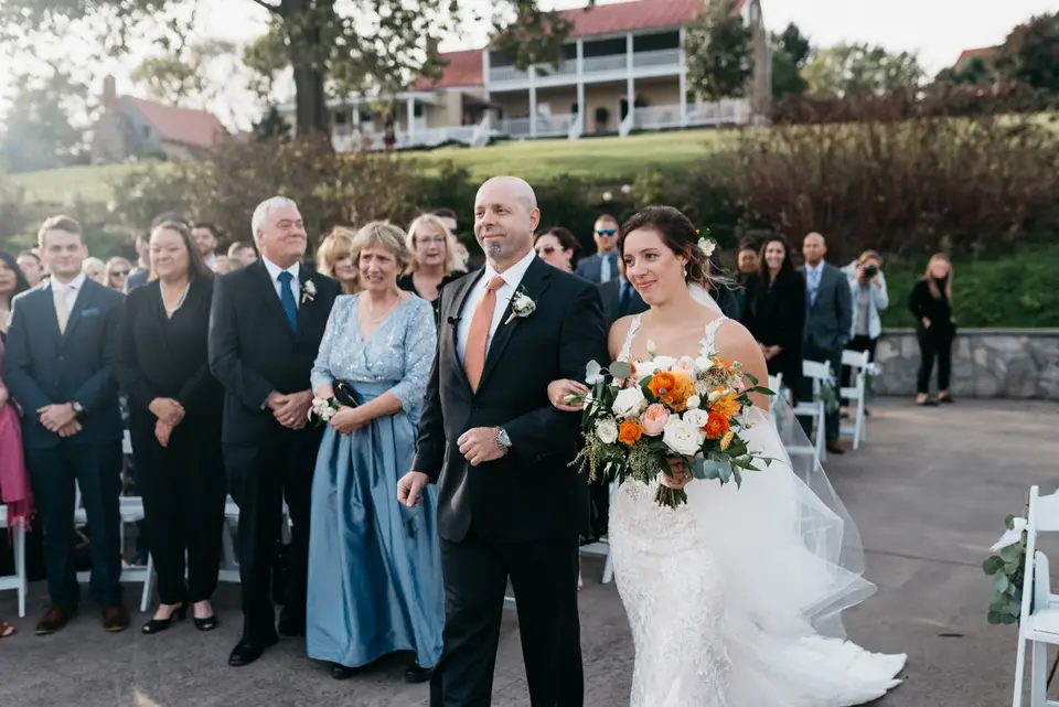 Bride in a lace gown with orange bouquet walking down the aisle on her father's arm past guests