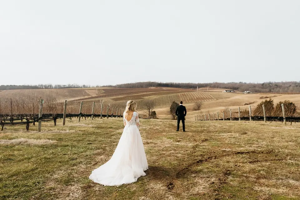 Bride in a white gown and groom standing apart in a vineyard in winter before a first look
