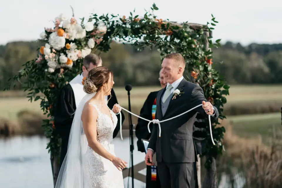 Couple tying a rope handfasting knot at an outdoor ceremony beneath a floral arch