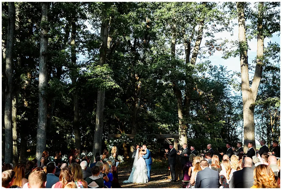 Outdoor wedding ceremony among tall trees as the couple kisses in front of seated guests