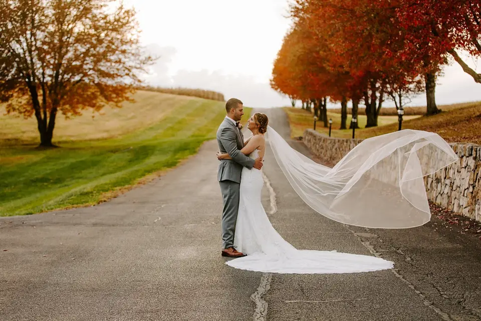 Bride and groom embracing on a country road lined with fall trees, her long veil blowing