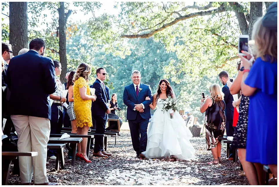 Father walking the bride down a wooded outdoor aisle past guests