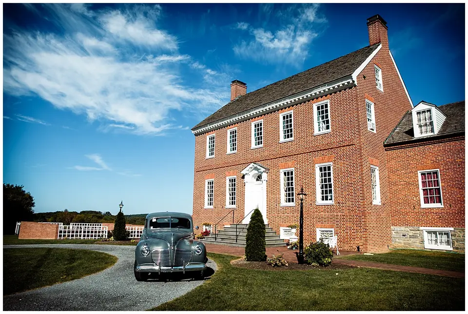 Vintage car parked in front of a red brick colonial-style house under a blue sky