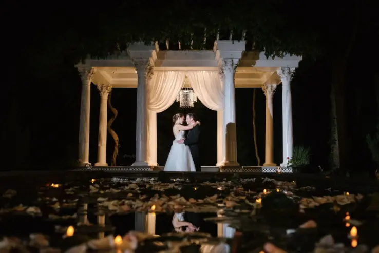 Bride and groom embracing in a lit white gazebo at night above a reflecting pool with candles