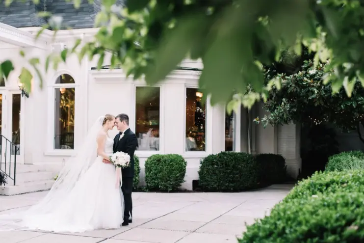 Bride and groom touching foreheads in front of a white building with hedges