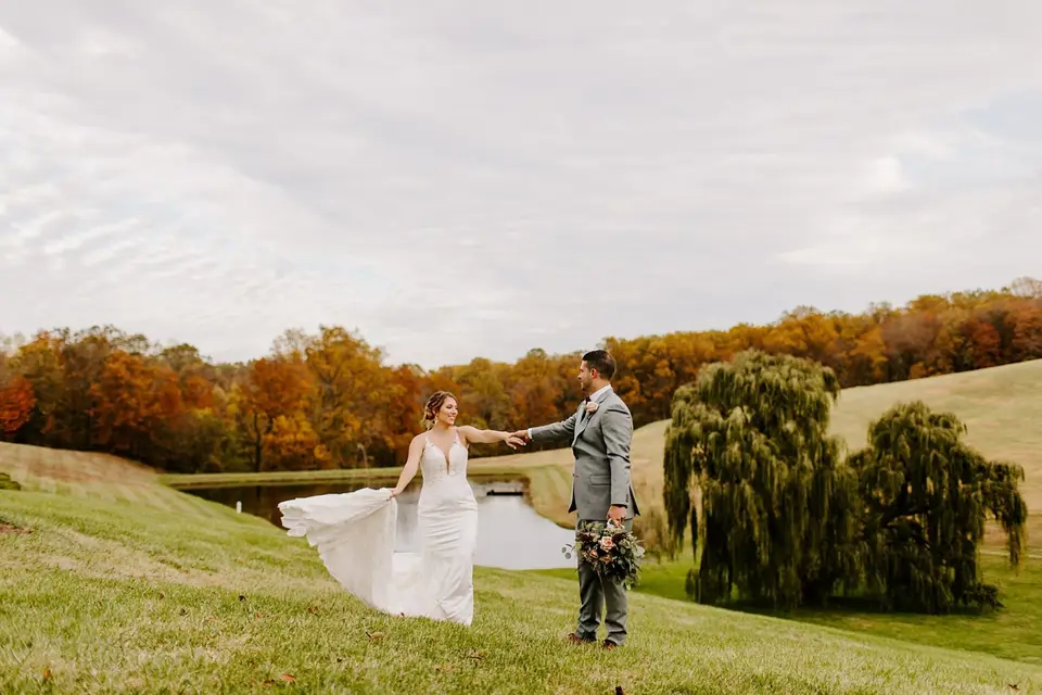 Bride and groom dancing apart on a grassy hill beside a pond with autumn foliage
