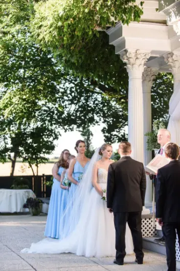 Bride and groom at an outdoor ceremony near white columns as the officiant reads and bridesmaids watch