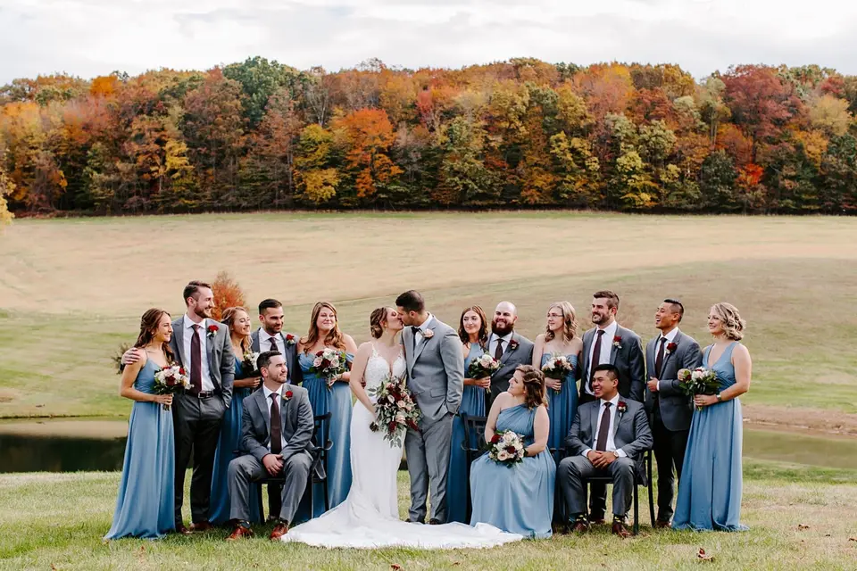 Wedding party in blue dresses and gray suits laughing around the couple with autumn trees behind