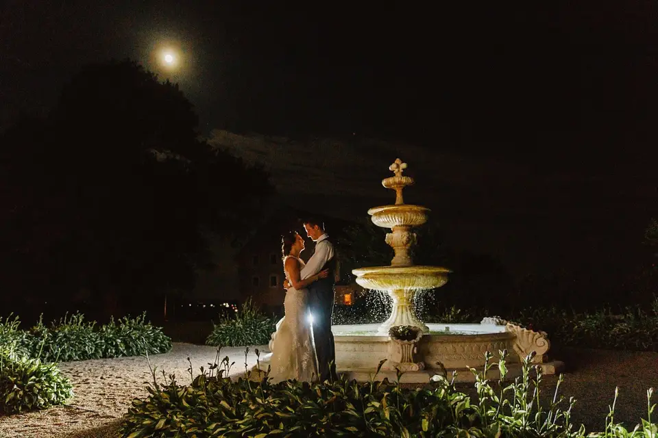 Couple embracing beside a lit stone fountain at night with the moon above