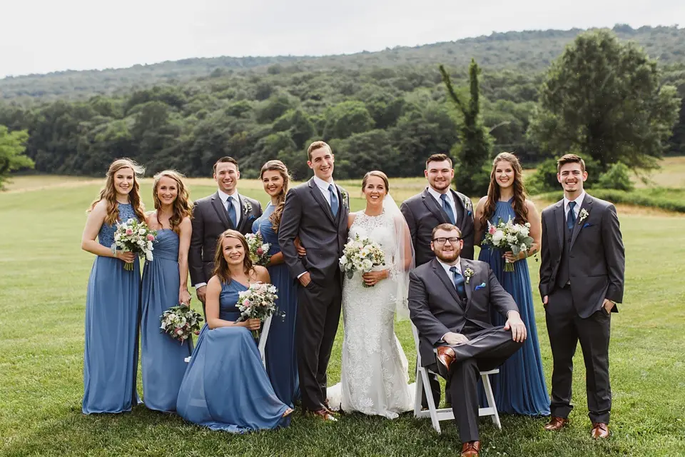 Bridal party in blue dresses and dark suits posed on a lawn with the couple, one man seated on a chair