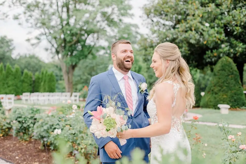 Groom in a blue suit laughing with the bride holding a bouquet in a garden