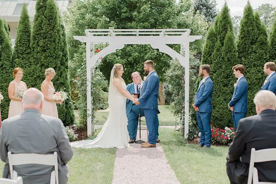 Couple exchanging vows under a white pergola as guests and groomsmen look on
