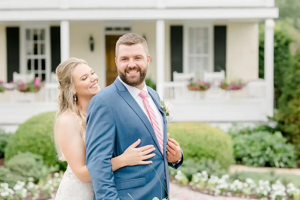 Groom in a blue suit smiling as the bride hugs him from behind in front of a porch