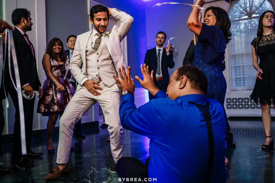 Groom in a cream suit dances while a guest in blue kneels and claps in front of him