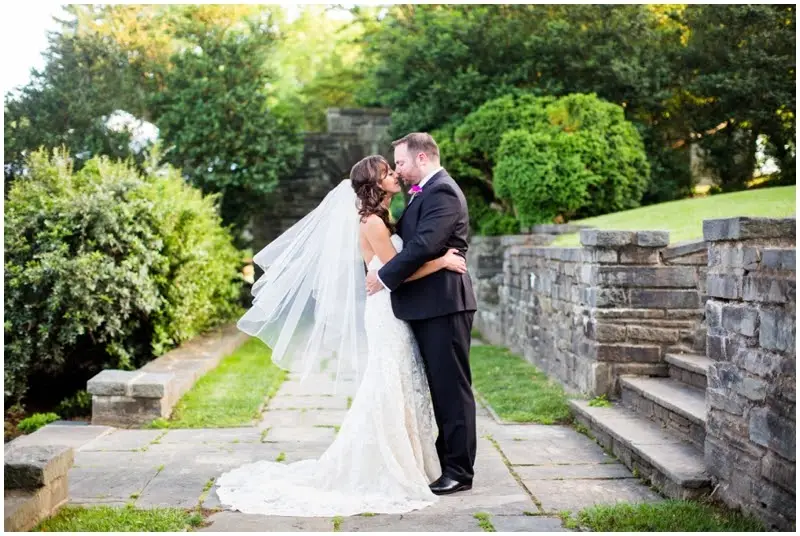 Bride and groom kiss on a stone path beside a stone wall and green hedges