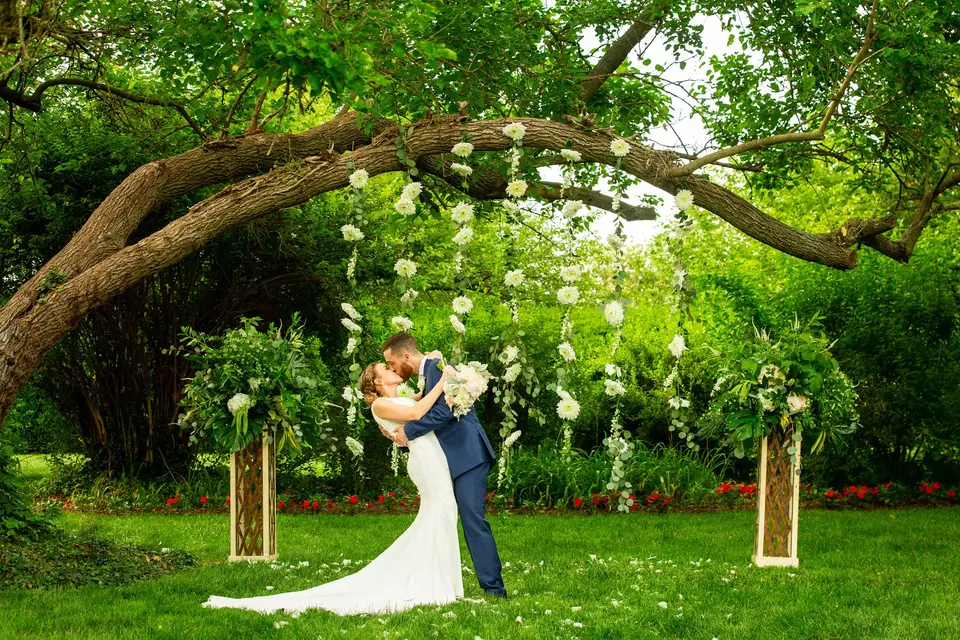 Bride and groom kissing under a branch hung with white flowers, flanked by floral arrangements