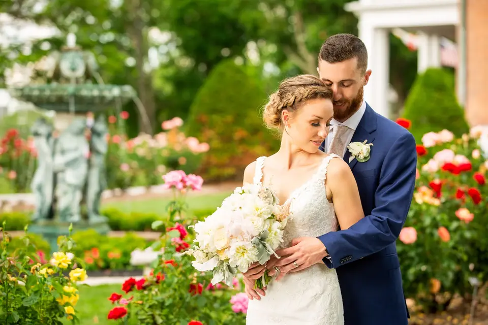 Groom embracing the bride from behind in a rose garden with a fountain statue behind them