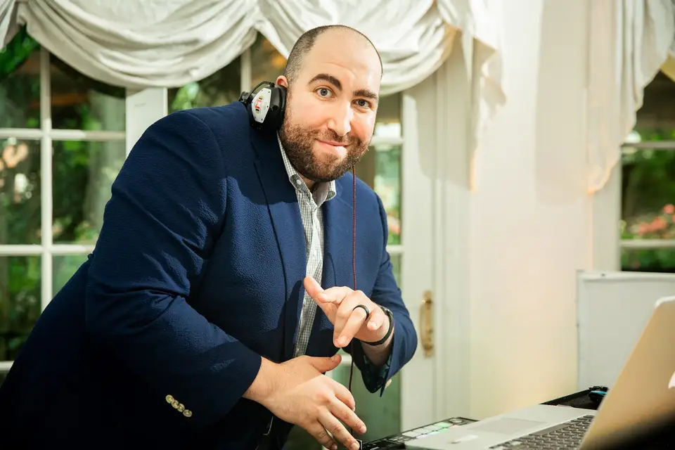 Smiling DJ in a navy blazer and headphones leaning over his laptop at an indoor wedding