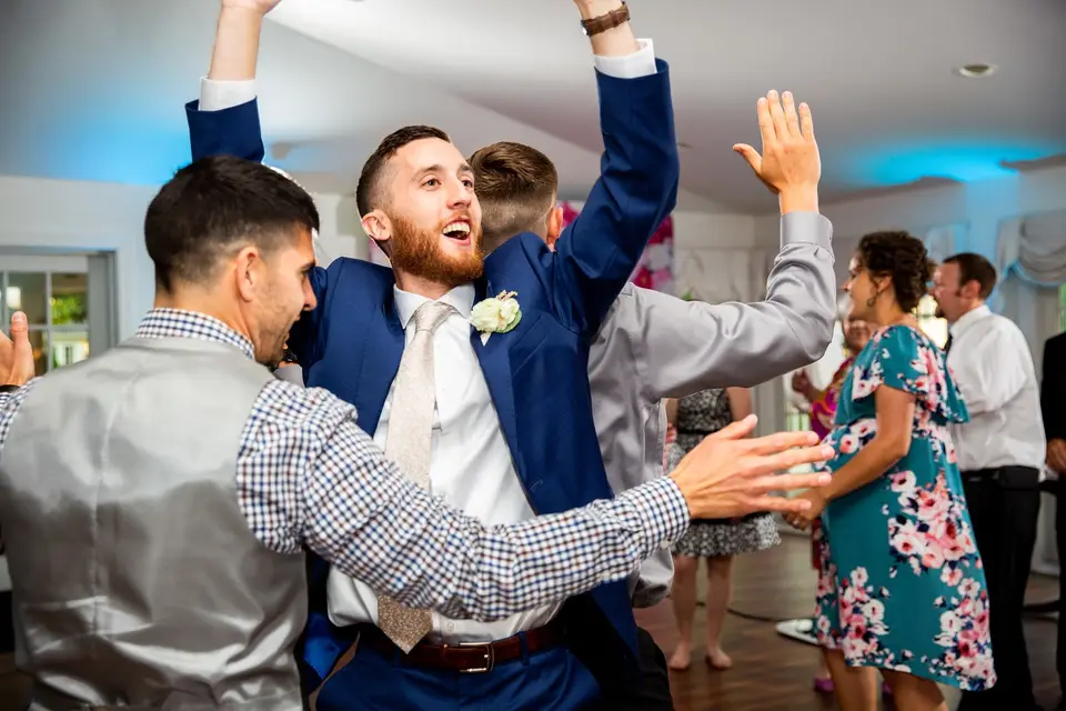 Groom in a blue suit with arms raised dancing with guests at a reception