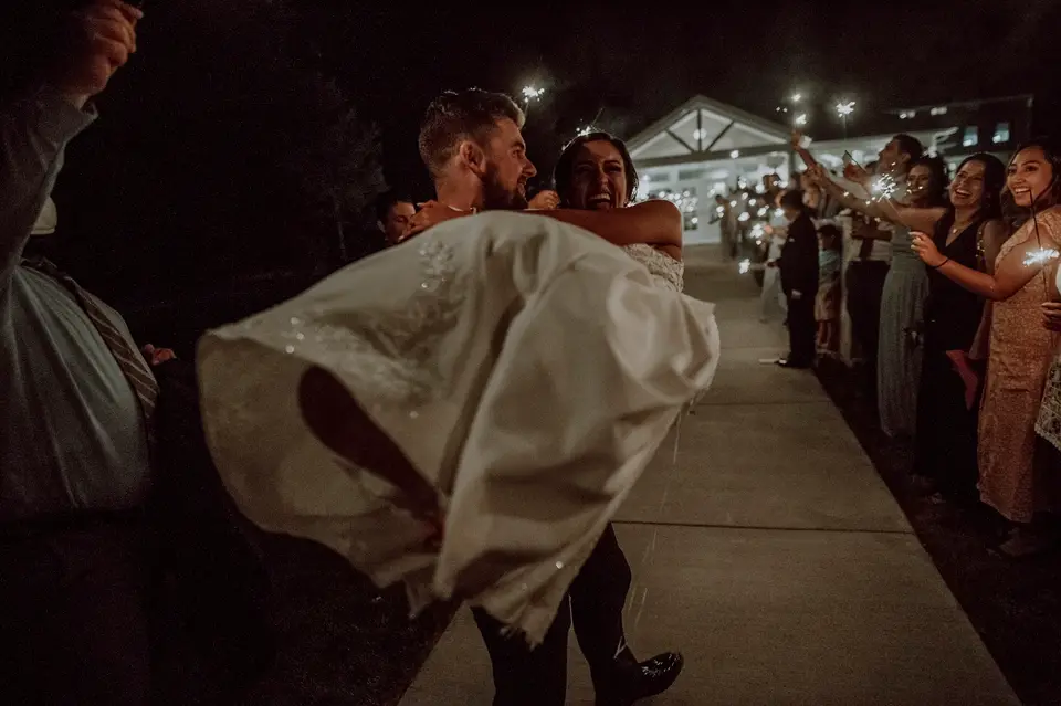 Groom carrying the bride through a sparkler send-off as guests line the path at night
