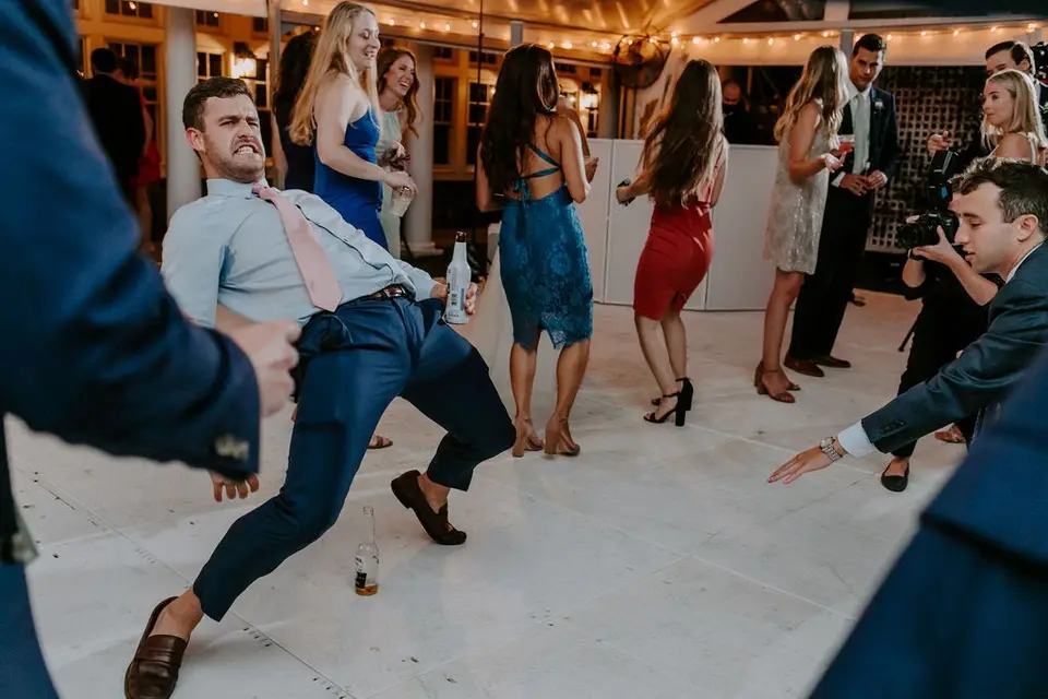 Guest in a blue shirt and pink tie leans back mid dance move on a white dance floor as others dance behind him