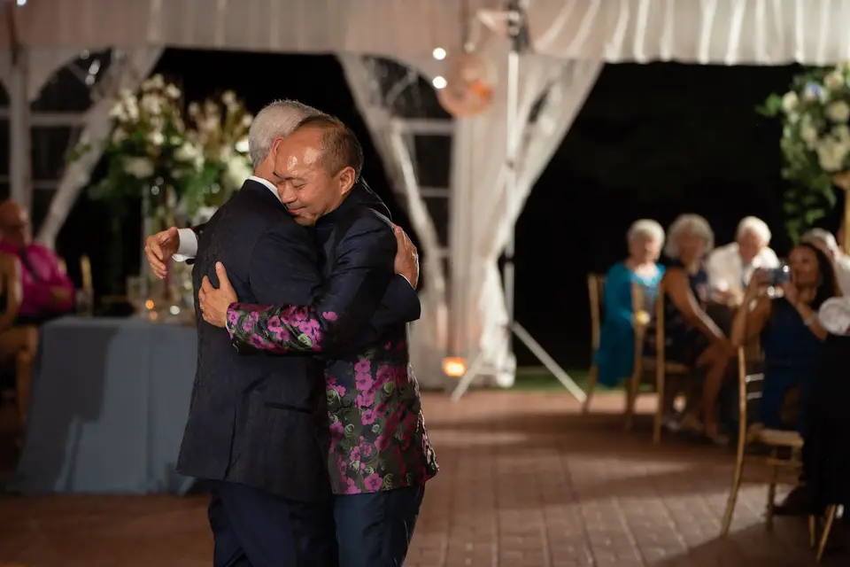 Two grooms hugging during a dance at an evening tent reception while guests watch from their seats