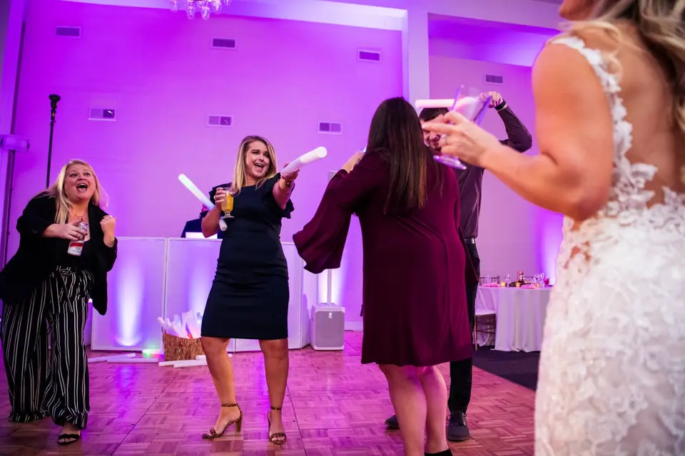 Guests dance with foam glow sticks on a parquet floor in a room lit with purple lights, bride's lace back in view