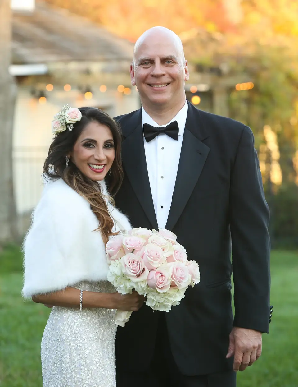 Smiling bride in a fur wrap and groom in a bow tie pose outdoors holding a pink rose bouquet