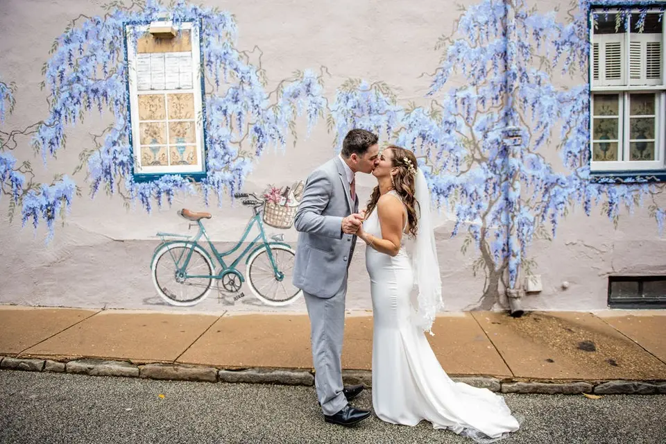 Bride and groom kissing on a street in front of a wall mural of wisteria and a bicycle