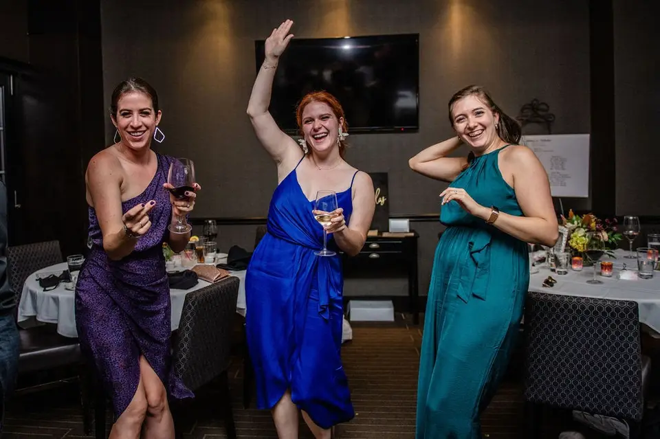 Three women in purple, blue and teal dresses laughing and dancing with drinks in hand at a reception