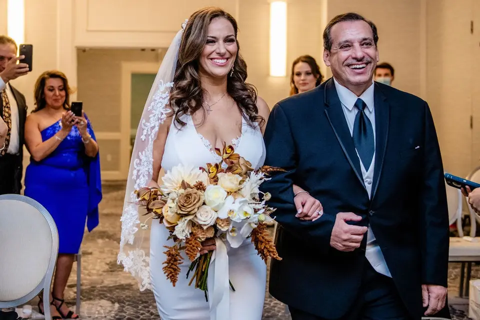 Smiling bride in a lace veil holding a bouquet, arm in arm with her father in a navy suit as guests take photos