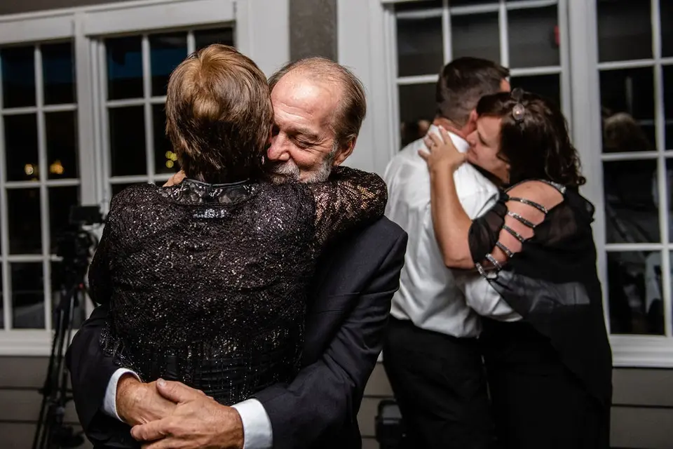 Older man and woman in an emotional hug at a wedding reception while two others embrace beside them