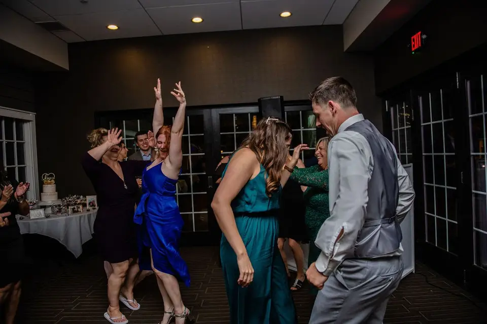 Guests dancing at a reception, a woman in a blue dress with arms raised and a man in a gray vest beside her
